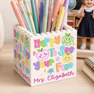 May include: A white square pencil holder constructed from interlocking building blocks. The exterior features colorful text reading Happy To See your Face, illustrated with smiling apple icons and flowers. The name Mrs. Elizabeth is printed at the base.