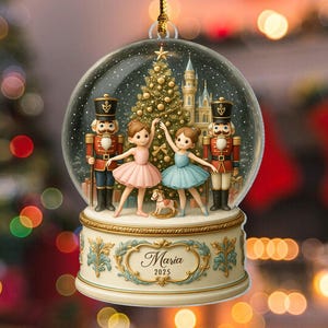 May include: A snow globe Christmas ornament with a gold hanging string. Inside, a Christmas tree, a castle, and two ballerinas in pink and blue tutus are visible. Two toy soldier nutcrackers stand on either side. The base reads "Maria 2025".