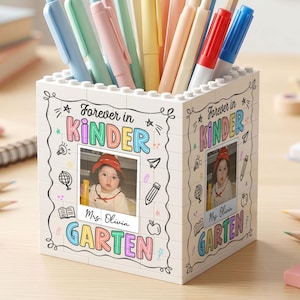 May include: A white square pencil holder constructed from building blocks, featuring a photo of a child and the text "Forever in KINDERGARTEN" with "Mrs. Olivia" printed below. The holder is filled with various colored pens and pencils.