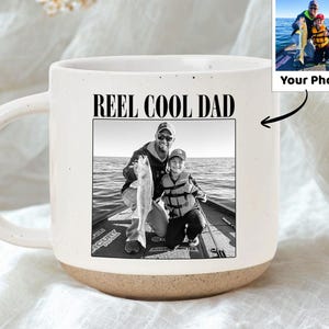 May include: White ceramic mug with a beige base and a large handle. The mug features a black and white photo of a father and child fishing, with the text "REEL COOL DAD" above the photo. A smaller photo is also included.