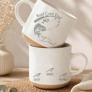 May include: Two white ceramic mugs with a tan base. One mug has "Reel Cool Dad" and a fish graphic. The other mug has three fish graphics with names below. A fishing-themed gift.