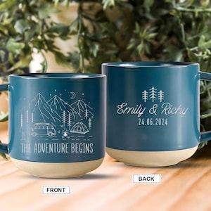 May include: Two teal blue ceramic mugs with white designs. The front mug has a mountain range, a camper van, a campfire, and a tent with the text "The Adventure Begins". The back mug has a line of trees and the text "Emily & Richy 24.06.2024".