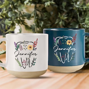 May include: Two ceramic coffee mugs with a tooth design and floral accents. The mugs are personalized with the name "Jennifer". One mug is white with a brown base and the other is teal with a brown base.