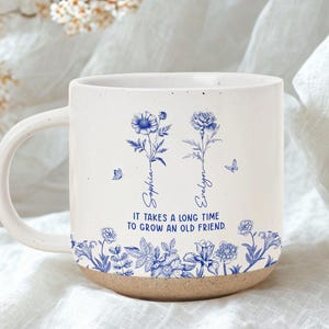May include: White ceramic mug with a blue floral design and the names "Sophia" and "Evelyn." The mug features the quote "It takes a long time to grow an old friend." The bottom of the mug is a natural, unglazed colour.
