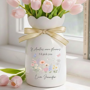 May include: A white ceramic vase with a scalloped rim, tied with a gold ribbon, holds pink tulips. The vase is decorated with a floral design and the text "If Aunties were flowers, I'd pick you, Love Jennifer."
