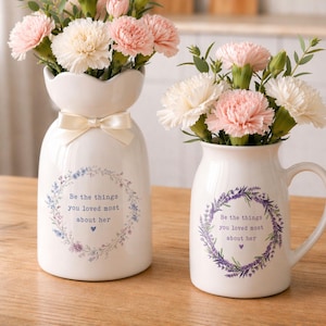 Peut inclure: Deux vases en c&eacute;ramique blanche avec des arrangements floraux. L'un a un n&oelig;ud, l'autre une anse. Les deux portent l'inscription "Be the things you loved most about her" dans une couronne florale. Les fleurs sont roses et blanches.