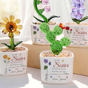 May include: A collection of glass flower sculptures in white pots. The flowers include a sunflower, tulips, a cactus, and lavender. Each pot has the text "TO MY Sister" and a personalized message. The pots are decorated with floral and butterfly accents.
