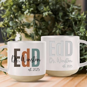 May include: Two white ceramic mugs with the text "EdD Dr. Winston est. 2025" on them. One mug has the text in colorful letters, while the other mug has the text in white letters.