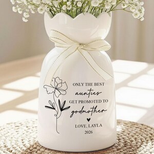 May include: A white ceramic vase with a scalloped rim, tied with a cream-colored ribbon, and filled with white flowers. The vase has a black floral design and the text "ONLY THE BEST aunties GET PROMOTED TO godmother LOVE, LAYLA 2026."