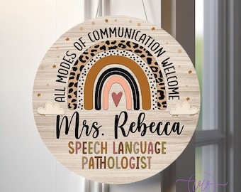 May include: A round wooden sign with a rainbow design featuring a leopard print pattern. The sign reads "All Modes of Communication Welcome" and "Mrs. Rebecca Speech Language Pathologist".
