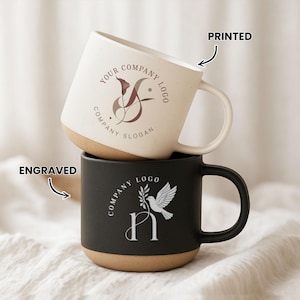 May include: A stacked pair of ceramic mugs featuring a cream-coloured top mug with a printed floral logo and text reading YOUR COMPANY LOGO COMPANY SLOGAN, and a black bottom mug with an engraved dove and leaf logo and text COMPANY LOGO.