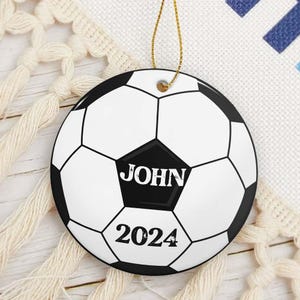 May include: A round soccer ball ornament with a gold string for hanging. The ornament is white with black pentagons and hexagons. The name "JOHN" is printed in white on a black pentagon, with "2024" below it.