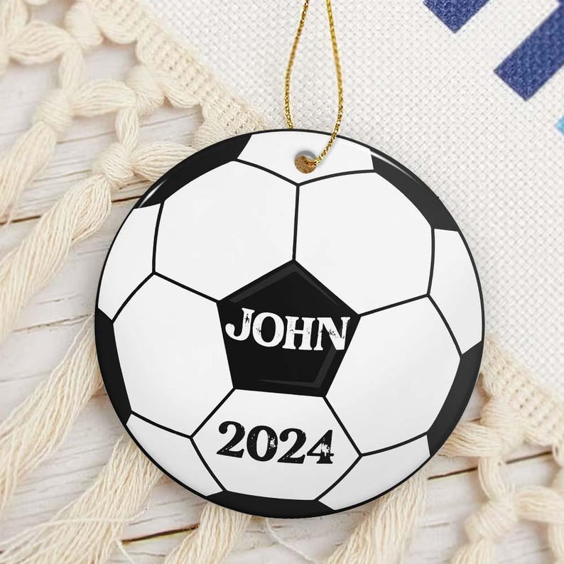 May include: A round soccer ball ornament with a gold string for hanging. The ornament is white with black pentagons and hexagons. The name "JOHN" is printed in white on a black pentagon, with "2024" below it.