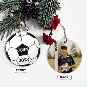 May include: A soccer-themed Christmas ornament. One side features a soccer ball design with the name "JOHN" and the year "2024". The other side displays a photo of a young man holding a soccer ball. The ornament hangs from a gold string.