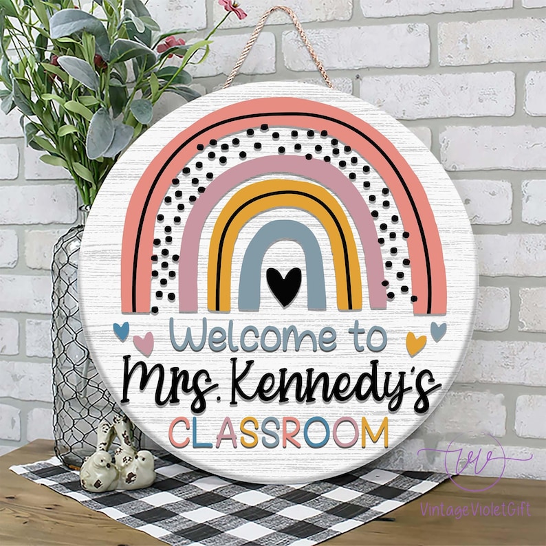 Pu&ograve; includere: Un cartello rotondo in legno con un design arcobaleno in rosa, giallo, blu e grigio. Il cartello dice "Benvenuti nella classe della Sig.ra Kennedy" in testo nero.