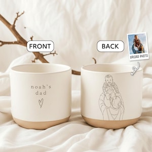 May include: A white ceramic mug with a tan base. The front of the mug has the text "noah's dad" and a heart symbol. The back of the mug features a line drawing of a person holding a child. A small photo is shown with the words "UPLOAD PHOTO".