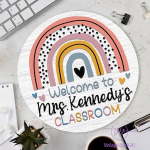 Pu&ograve; includere: Un cartello rotondo in legno con un design arcobaleno in rosa, giallo, blu e grigio con pois neri. Il cartello dice "Benvenuti nella classe della Sig.ra Kennedy" in testo nero.