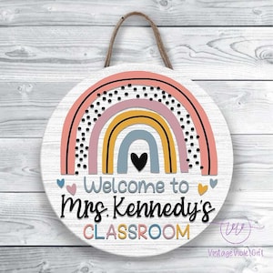 Pu&ograve; includere: Un cartello rotondo in legno bianco con un design arcobaleno in rosa, giallo, blu e grigio. L'arcobaleno ha pois neri. Il cartello dice "Benvenuti nella classe della Sig.ra Kennedy".