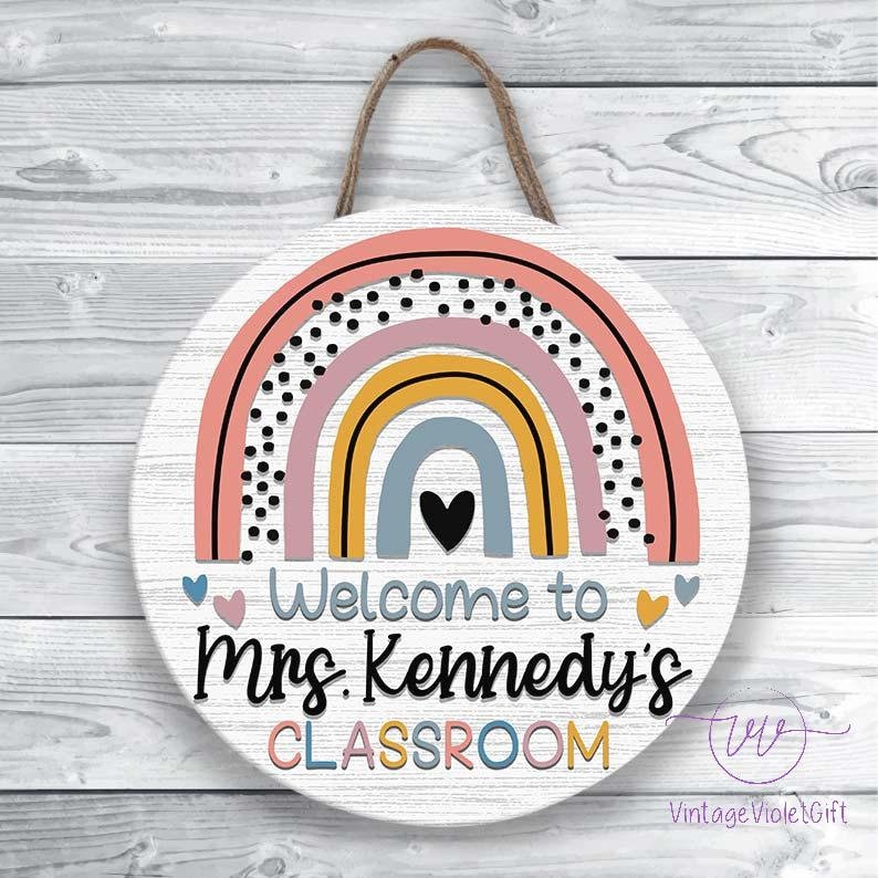 Pu&ograve; includere: Un cartello rotondo in legno bianco con un design arcobaleno in rosa, giallo, blu e grigio. L'arcobaleno ha pois neri. Il cartello dice "Benvenuti nella classe della Sig.ra Kennedy".