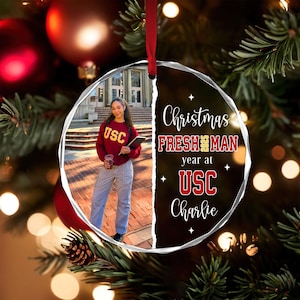 May include: A clear, round Christmas ornament with a photo of a person wearing a USC sweatshirt. The ornament has the text "Christmas, Freshman year at USC, Charlie". The ornament hangs from a red ribbon.