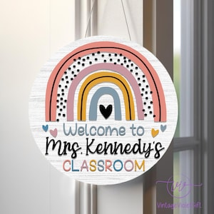 Pu&ograve; includere: Un cartello rotondo in legno con un design a arcobaleno e il testo "Welcome to Mrs. Kennedy's CLASSROOM". L'arcobaleno &egrave; fatto di strisce rosa, gialle, blu e grigie con pois neri sulla striscia rosa. Ci sono piccoli cuori rosa su entrambi i lati del testo.