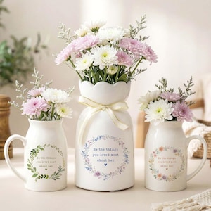Peut inclure: Trois vases en c&eacute;ramique blanche avec des arrangements floraux. Les vases sont d&eacute;cor&eacute;s d'une couronne florale et du texte "Be the things you loved most about her". Les fleurs sont blanches et roses.