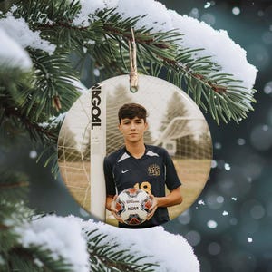 May include: A round ceramic ornament featuring a young man in a soccer uniform holding a soccer ball. The ornament has a string for hanging and is set against a snowy evergreen backdrop. The text "N GOAL" is visible.