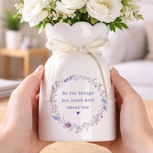 Peut inclure: Un vase en c&eacute;ramique blanc avec un bord festonn&eacute;, nou&eacute; avec un ruban cr&egrave;me, contenant des roses blanches et de la verdure. Le vase porte une couronne florale et le texte "Be the things you loved most about her".