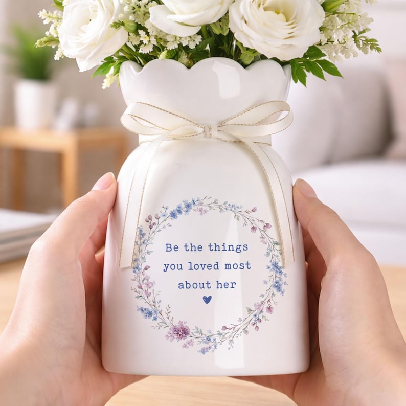 Peut inclure: Un vase en c&eacute;ramique blanc avec un bord festonn&eacute;, nou&eacute; avec un ruban cr&egrave;me, contenant des roses blanches et de la verdure. Le vase porte une couronne florale et le texte "Be the things you loved most about her".
