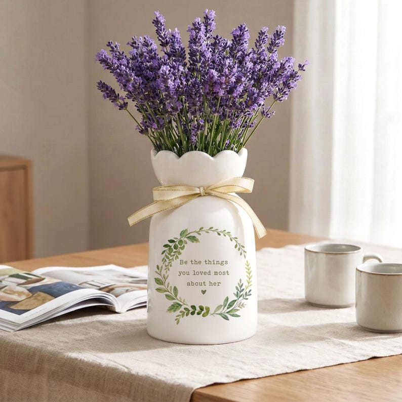 Peut inclure: Un vase en c&eacute;ramique blanc &agrave; bord festonn&eacute; contient un bouquet de lavande violette. Le vase est orn&eacute; d'un ruban dor&eacute; et d'une couronne verte avec le texte "Be the things you loved most about her."