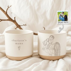 May include: Two off-white ceramic mugs with speckled textures. One mug features the text "charlotte's mama" above a small heart outline. The other mug displays a dark brown line art illustration of a person lifting a small child into the air.