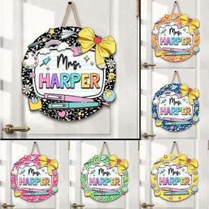 May include: Round wooden teacher door sign with a black and white composition notebook design. The sign features the name "Mrs. Harper" in colorful letters, a yellow bow, and school-themed illustrations. It hangs from a natural rope.