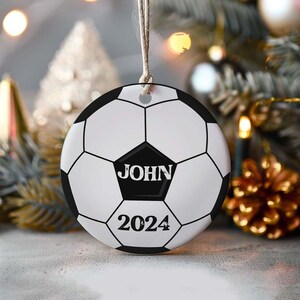May include: A round Christmas ornament designed as a soccer ball, featuring black and white pentagons and hexagons. The name "JOHN" is printed in bold white letters on a black pentagon, with "2024" below it. The ornament hangs from a string.