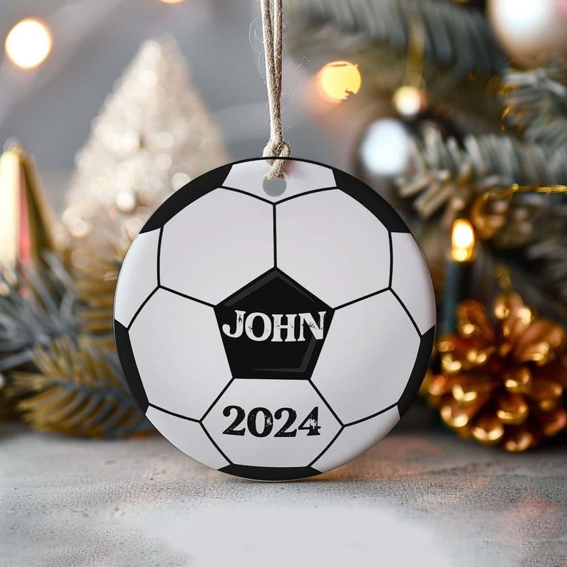 May include: A round Christmas ornament designed as a soccer ball, featuring black and white pentagons and hexagons. The name "JOHN" is printed in bold white letters on a black pentagon, with "2024" below it. The ornament hangs from a string.