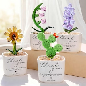 May include: A collection of glass flower sculptures in white ceramic pots. The flowers include a yellow sunflower, pink tulips, a green cactus, and a purple hyacinth. Each pot has the words "thank you" and a message written on it.
