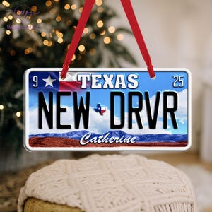 May include: A rectangular ornament resembling a Texas license plate, featuring the text "TEXAS" and "NEW DRVR". The ornament has a blue sky background with a Texas flag and a landscape scene at the bottom. A red ribbon is attached for hanging.