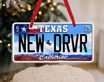 May include: A rectangular ornament resembling a Texas license plate, featuring the text "TEXAS" and "NEW DRVR". The ornament has a blue sky background with a Texas flag and a landscape scene at the bottom. A red ribbon is attached for hanging.