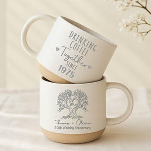 May include: Two stacked white ceramic mugs with beige bases and white handles. The top mug reads "DRINKING COFFEE Together SINCE 1975". The bottom mug features a tree design with a heart and the text "Thomas + Olivia 50th Wedding Anniversary".