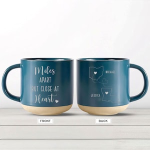 May include: A teal blue ceramic mug with a white inscription that reads "Miles Apart But Close At Heart" with a small heart symbol. The back of the mug features a map of Ohio and Louisiana with the names "Michael" and "Jessica" and a dotted line connecting the two states.