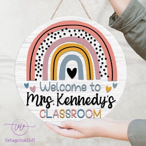 Pu&ograve; includere: Un cartello rotondo in legno con un design arcobaleno in tonalit&agrave; di rosa, giallo, blu e grigio. L'arcobaleno ha pois neri e un cuore nero al centro. Il cartello dice "Benvenuti nella classe della Sig.ra Kennedy" in testo nero.