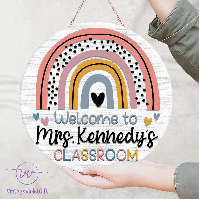 Pu&ograve; includere: Un cartello rotondo in legno con un design arcobaleno in tonalit&agrave; di rosa, giallo, blu e grigio. L'arcobaleno ha pois neri e un cuore nero al centro. Il cartello dice "Benvenuti nella classe della Sig.ra Kennedy" in testo nero.