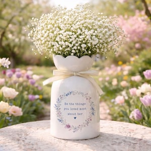 Peut inclure: Un vase en c&eacute;ramique blanc avec un dessus festonn&eacute;, rempli de fleurs de gypsophile blanches. Le vase a un ruban cr&egrave;me et un motif de couronne florale avec le texte "Be the things you loved most about her."