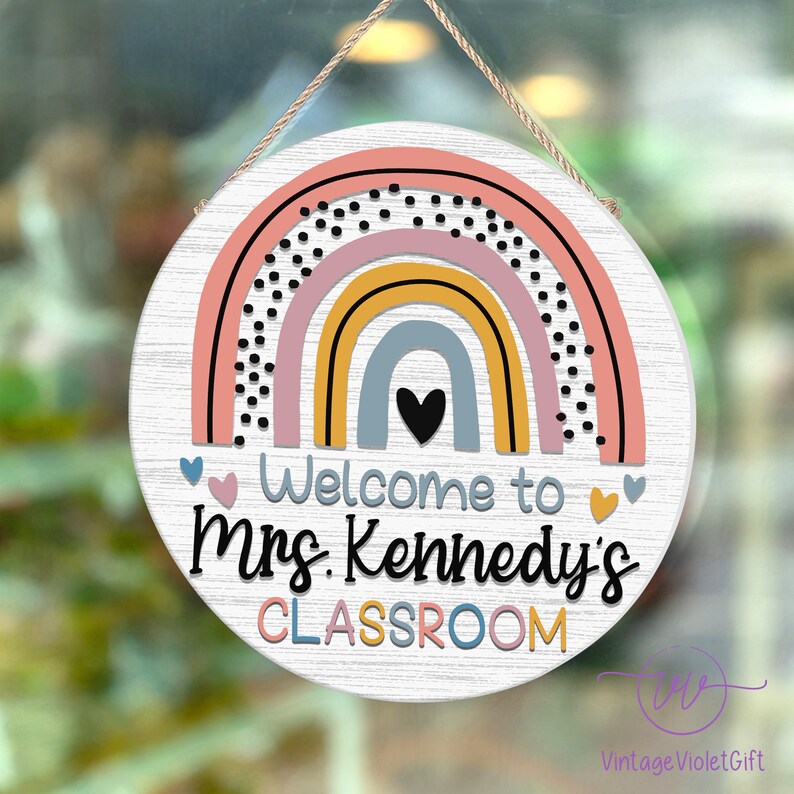 Pu&ograve; includere: Un cartello rotondo in legno con un design arcobaleno in rosa, giallo, blu e grigio con pois neri. Il cartello dice "Benvenuti nella classe della Sig.ra Kennedy" in testo nero.