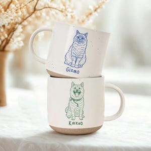May include: Two white ceramic mugs stacked on top of each other. The top mug has a blue illustration of a cat and the name "Gizmo". The bottom mug has a green illustration of a dog and the name "Kazuo". Both mugs have a speckled design and a tan base.