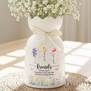 May include: White ceramic vase with a scalloped rim and a cream-colored ribbon. The vase is decorated with floral illustrations, butterflies, and the word "friends" with a quote. The vase is filled with white flowers.