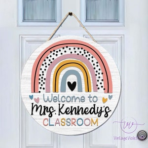 Pu&ograve; includere: Un appendiporta rotondo in legno con un design arcobaleno in rosa, giallo, blu e grigio. L'arcobaleno ha pois neri. Il testo "Welcome to Mrs. Kennedy's CLASSROOM" &egrave; scritto in bianco.