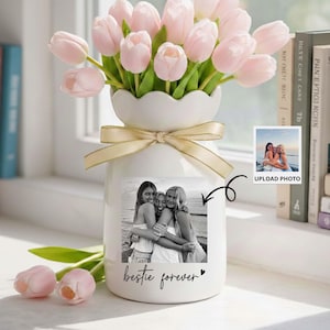 May include: A white ceramic vase with a scalloped top, tied with a gold ribbon, holds pink tulips. A black and white photo of three women is affixed to the vase, with the words "bestie forever" below. A small photo with the words "UPLOAD PHOTO" is also present.