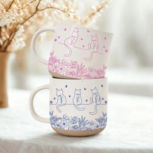 May include: Two stacked ceramic mugs with a white base and speckled beige bottom. The top mug has pink cat illustrations and floral designs, with names. The bottom mug features blue cat illustrations and floral designs, with names.