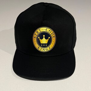 Op de afbeelding: Zwarte baseballcap met een platte klep. De cap heeft een goud en marineblauw embleem op de voorkant met de tekst "STRT CODE KINGZ" rond een kroon en drie sterren. De cap is gemaakt van een stevige stof.