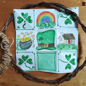 Può includere: Un'opera d'arte artigianale a tema San Patrizio con una griglia di nove quadrati con illustrazioni ad acquerello. L'opera comprende un arcobaleno, trifogli, un vaso d'oro, un cappello verde e una casa. L'opera è circondata da una ghirlanda intrecciata.